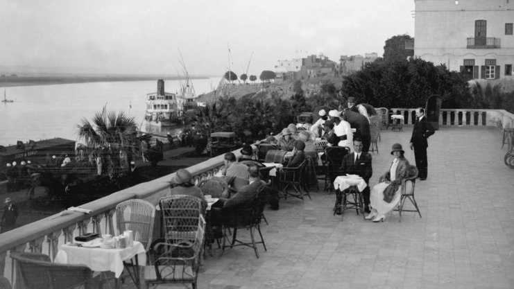 Photographie ancienne en noir et blanc de la terrasse du Winter Palace Hotel à Louxor, avec des touristes fortunés attablés pour le thé, surplombant le Nil et la rive ouest où se profile la Vallée des Rois, dans les années 1920.