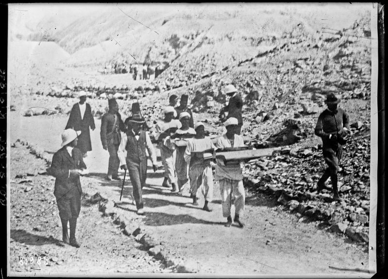Photographie d'époque en noir et blanc de l'archéologue Howard Carter supervisant le transport d'objets découverts dans le tombeau de Toutankhamon à Louxor, montrant des ouvriers égyptiens portant des corbeilles, des paniers et des caisses contenant des artefacts dont des pantoufles et des perles.