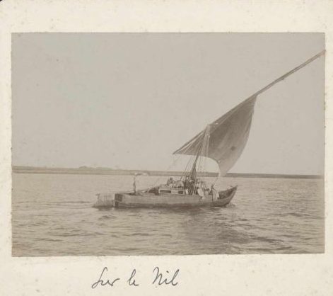 Photographie ancienne d'une felouque à voile sur le Nil, prise entre 1895 et 1899. L'embarcation traditionnelle glisse sur le fleuve, avec des rives désertiques en arrière-plan. Source : BNF.