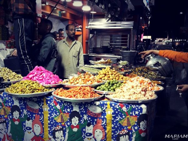 Restaurant de rue égyptien (comme un vendeur de koshari ou de grillades) bondé de clients attablés tard dans la nuit pendant le Ramadan, avec une ambiance vivante et des lumières vives.