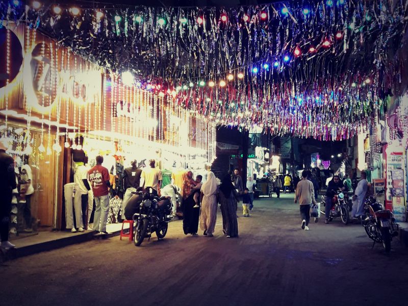 Rue commerçante égyptienne décorée pour le Ramadan avec une arche de fanions et de guirlandes lumineuses colorées traversant la chaussée, créant une ambiance festive.