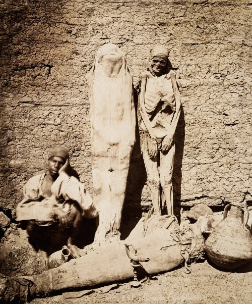 Photographie ancienne en noir et blanc d'un vendeur égyptien accroupi au milieu de momies et de fragments de sarcophages exposés sur un marché, vers 1877.
Photographie de Jean-Gilles Berizzi, vers 1877. © RMN-Grand Palais.