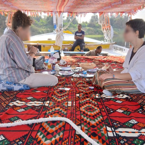 Intérieur d'une felouque aménagée pour le repas. Deux voyageurs sont attablés sur le pont, devant des plats locaux : salades, pain, et tajine. L'ambiance est chaleureuse et conviviale, typique des repas partagés avec l'équipage lors d'une croisière sur le Nil.