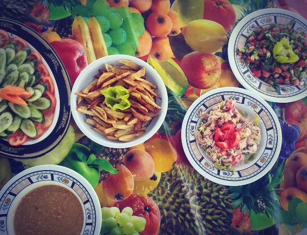 Table dressée sur le pont d'une felouque, recouverte de plats locaux : salades, pain frais, houmous, légumes cuisinés et tajine. La vaisselle simple et colorée repose sur une nappe, dans une lumière chaude typique d'un repas pris sur le Nil, au rythme du fleuve.
